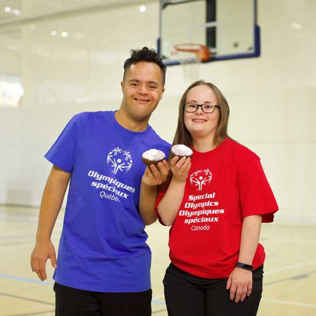 Deux athlètes sourient à la caméra. L'homme à gauche porte un t-shirt bleu des Olympiques spéciaux du Québec et la femme à droite porte un t-shirt rouge des Olympiques spéciaux du Canada. Tous deux sourient et tiennent un beignet. 