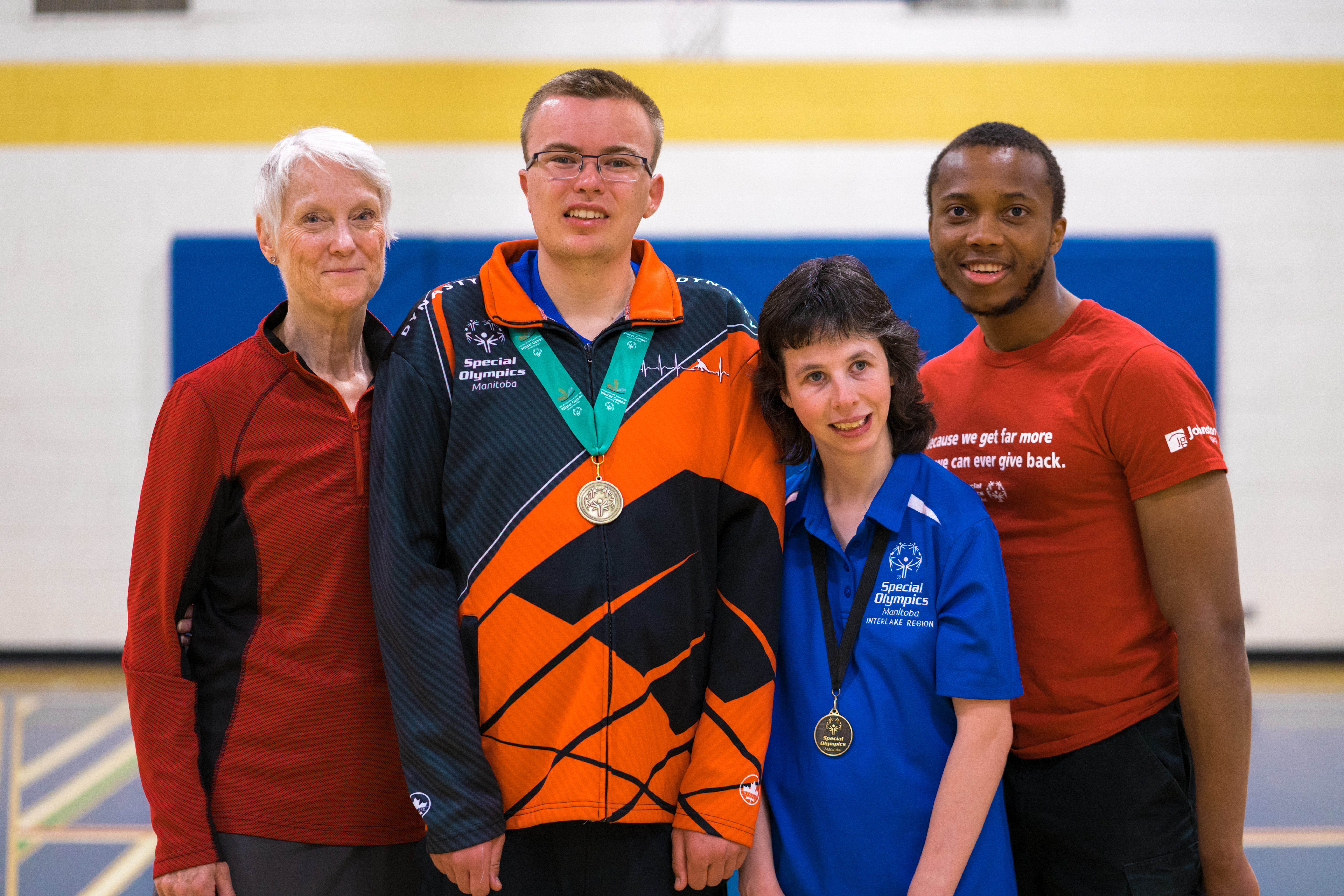Four adults standing together smiling at the camera. All are in athletic gear. Two have medals around their necks. Four adults standing together smiling at the camera. All are in athletic gear. Two have medals around their necks.