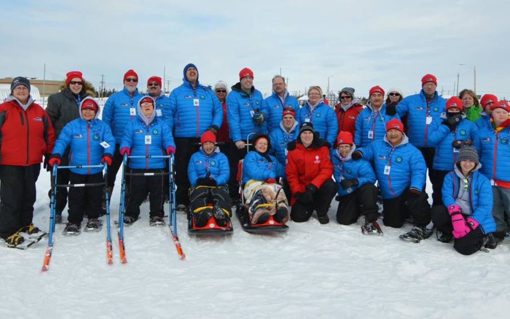 A large group photo of a Special Olympics Newfoundland & Labrador team. They are all wearing blue puffer jackets and some are additionally wearing red toques. It is wintertime and everyone is standing outside in the snow.