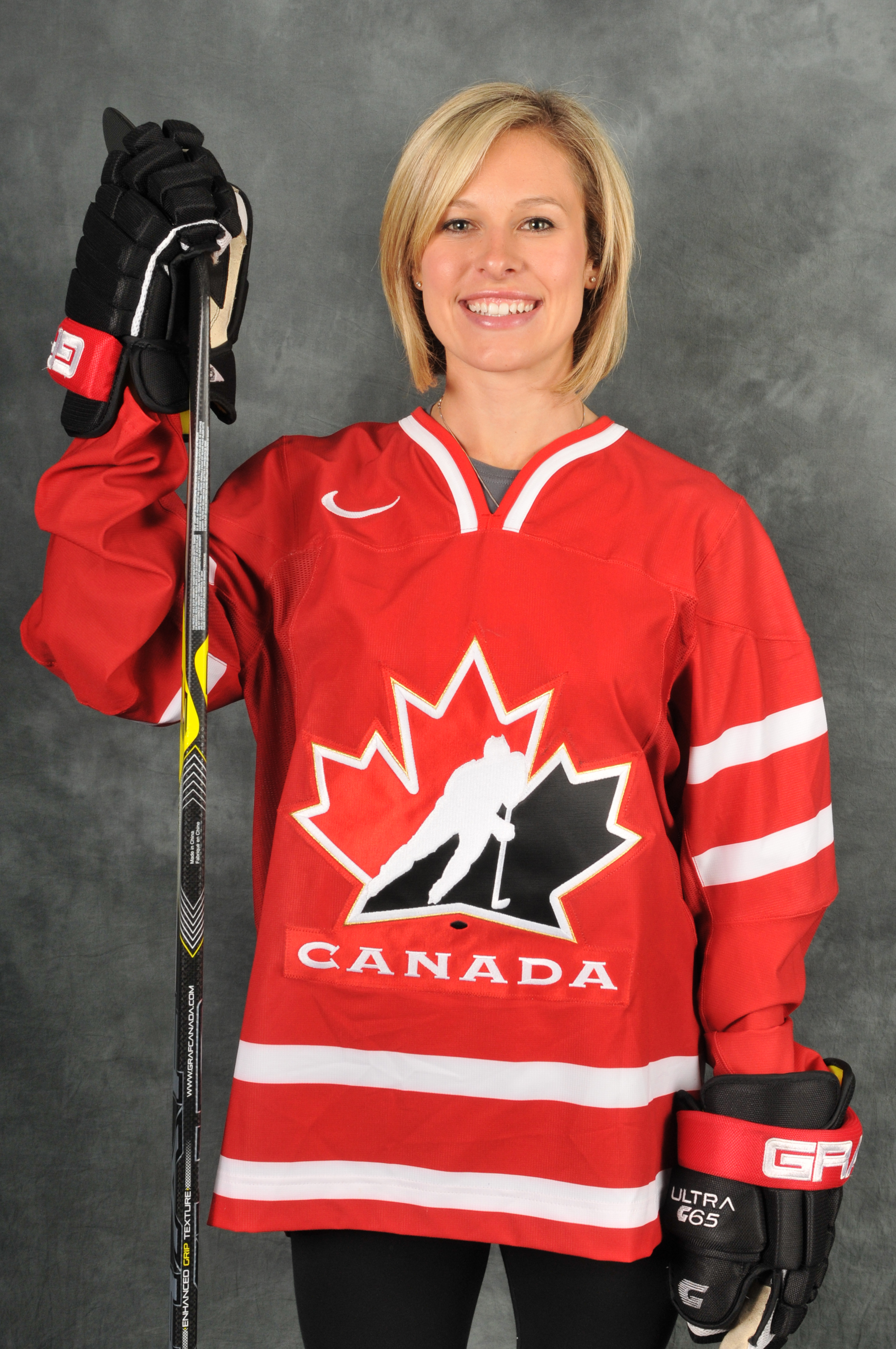 Headshot of a woman short blond hair wearing a Team Canada hockey jersey. She is wearing hockey gloves and holding a hockey stick. Headshot of a woman short blond hair wearing a Team Canada hockey jersey. She is wearing hockey gloves and holding a hockey stick.