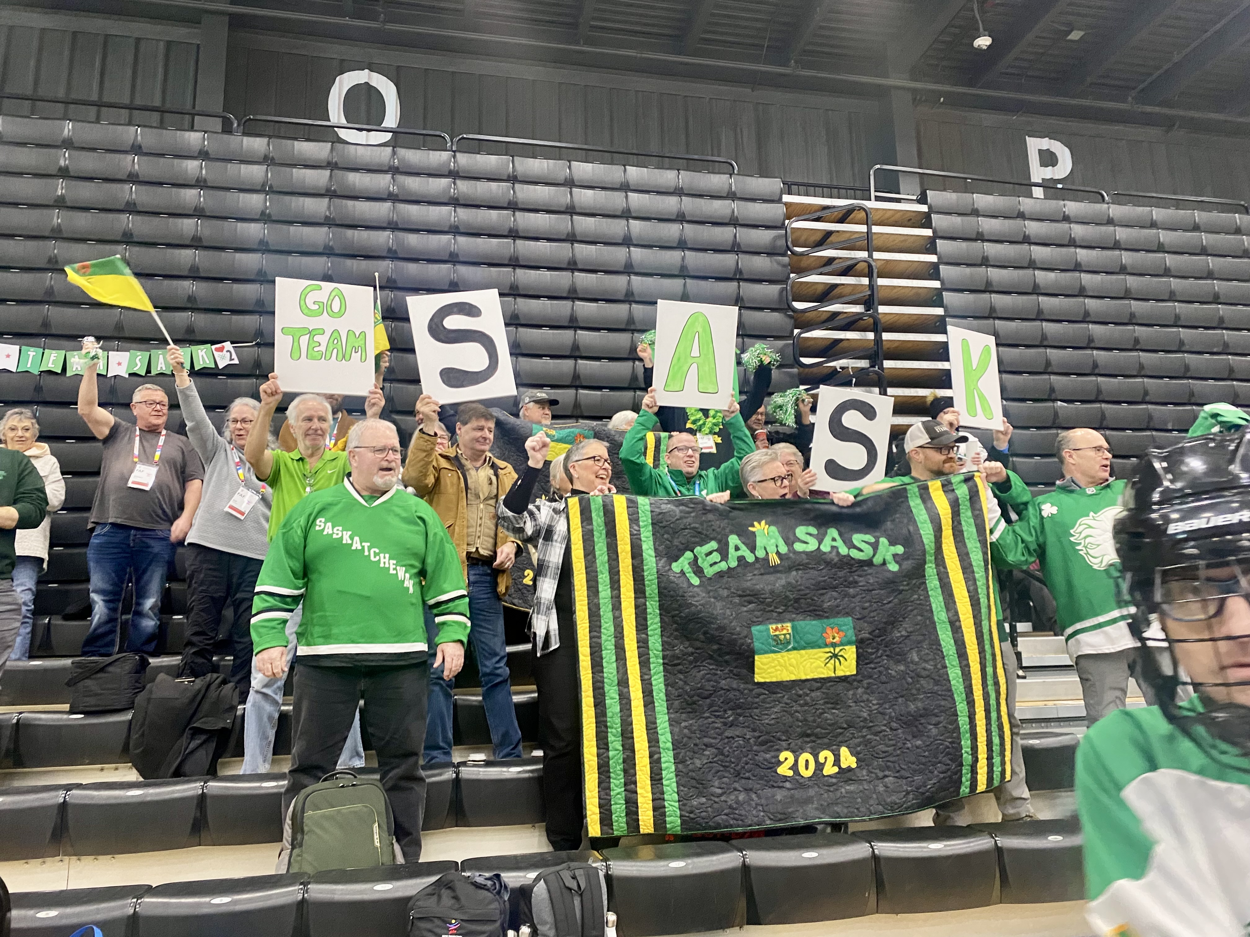 A crowd of people in green, are sitting on the bleachers cheering for Team Saskatchewan. They are holding up flags, banners, and poster boards showing support for the team. Everyone looks happy and are cheering the athletes on.