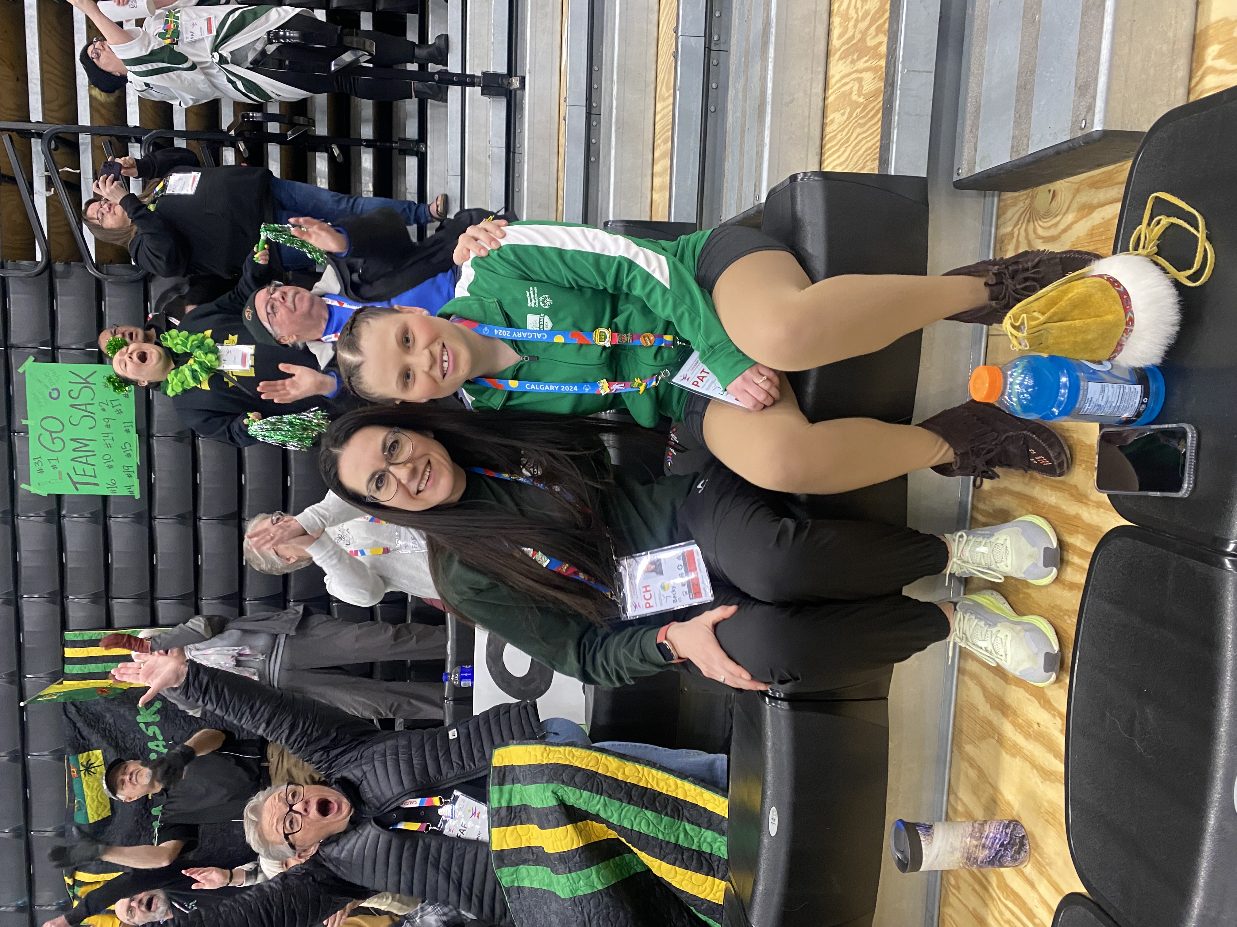 An athlete is sitting with her coach. Their arms are wrapped around behind each other. They are sitting in the bleachers of a sport event. The people behind them are all cheering.