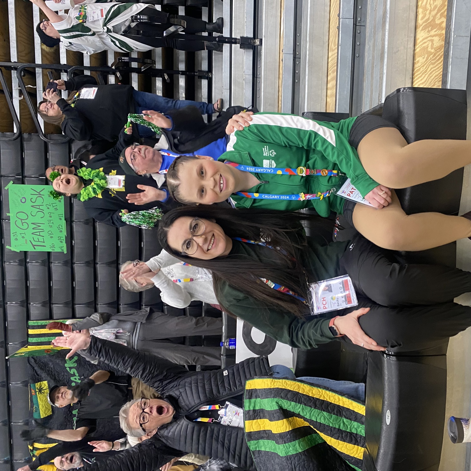 An athlete is sitting with her coach. Their arms are wrapped around behind each other. They are sitting in the bleachers of a sport event. The people behind them are all cheering.