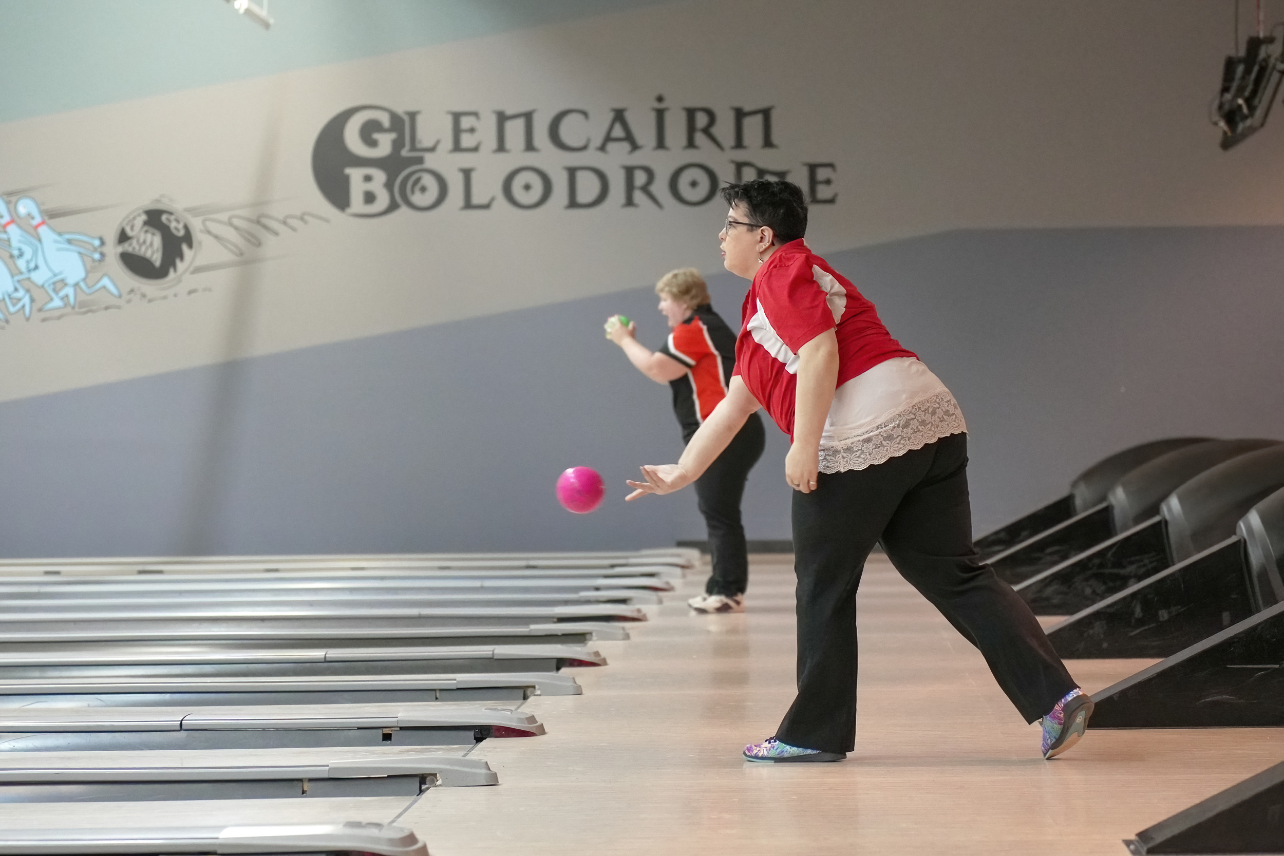 An action shot photo of a bowling athlete. The photo is taken from the side and you can see the start of many different bowling lanes. In the photo, two bowlers can be seen playing the game. The bowler closest to the camera is throwing a pink bowling ball which can be seen midair.