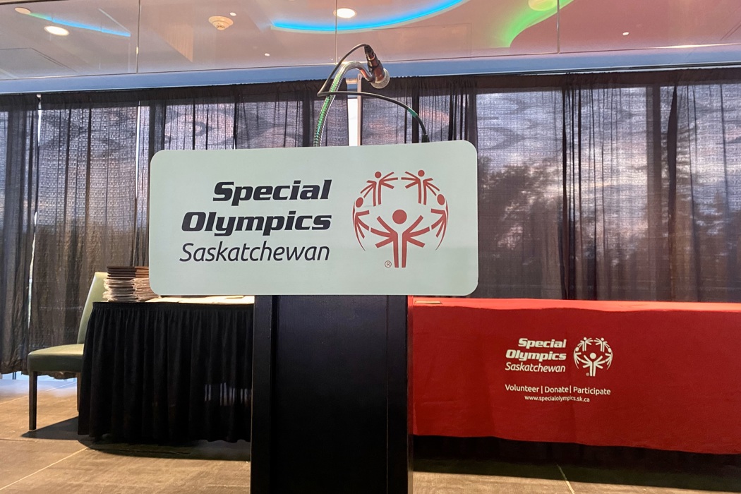 An image of a podium at a fancy event. The podium has a sign on it that reads, "Special Olympics Saskatchewan". Behind the podium is a long table in a red table cloth. The tablecloth also reads, "Special Olympics Saskatchewan".