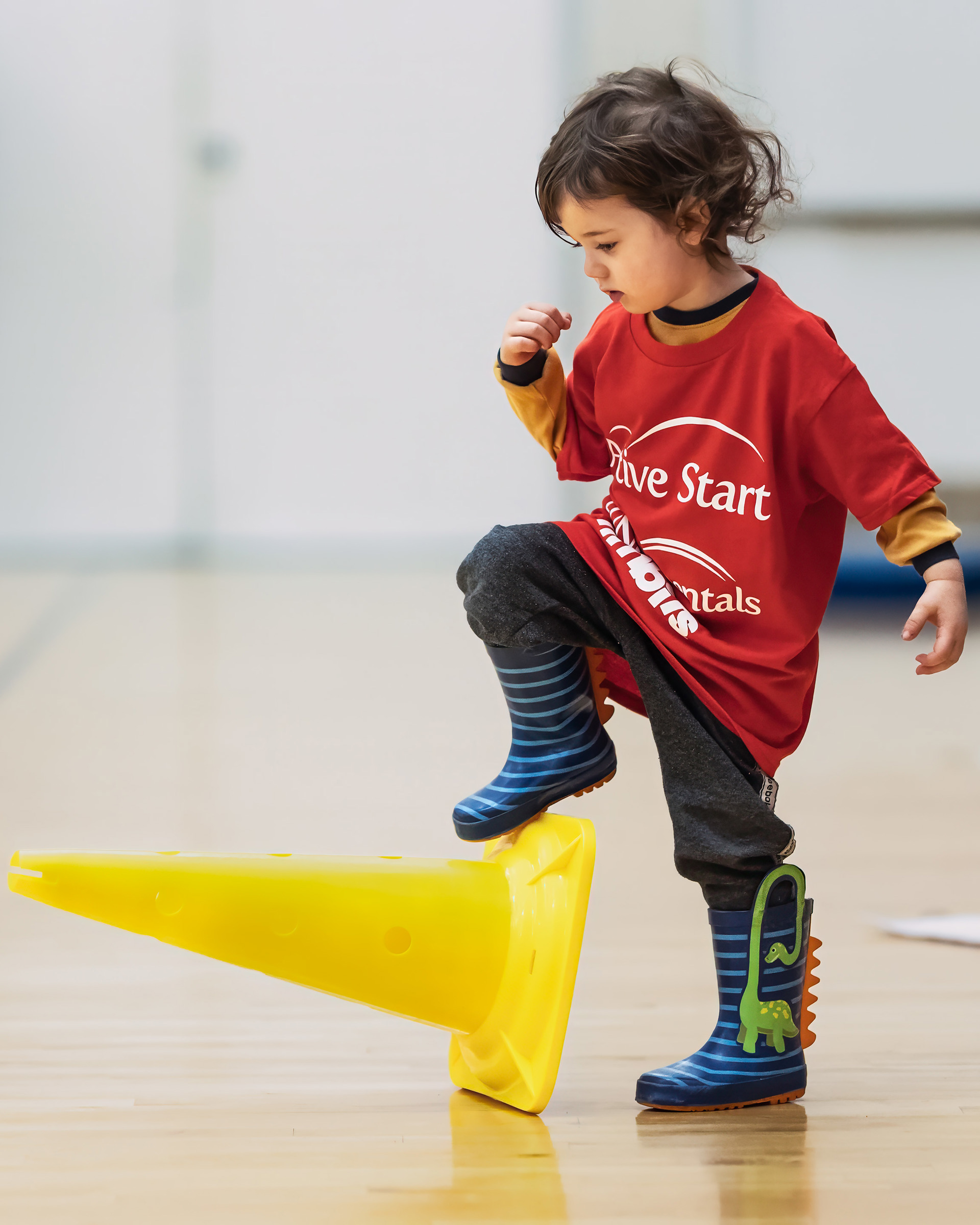 Petite fille en t-shirt rouge Active Start, bottes de pluie bleues à rayures, le pied posé sur un cône jaune couché sur le côté. En arrière-plan, un gymnase.
