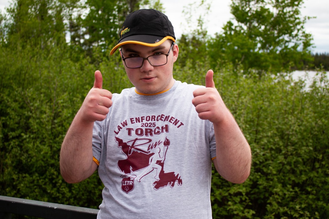 A half body photo of an athlete giving the camera two thumbs up. He is wearing a grey t-shirt that reads "Law Enforcement 2025 Torch Run" on it and a black baseball cap. Behind him are lots of greenery, trees, and bushes.