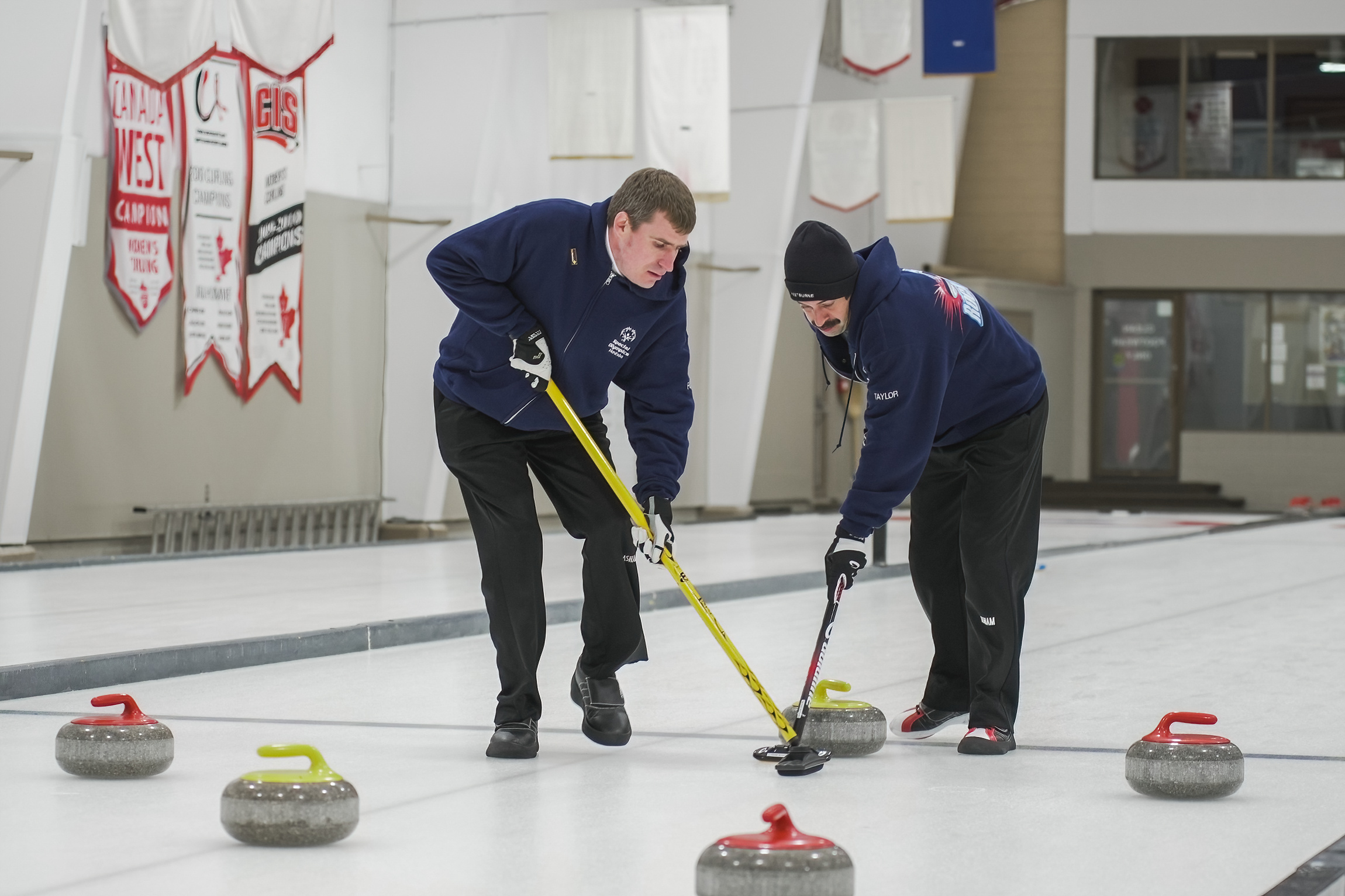 Two athletes participating in a curling match. There are three red and two yellow curling stones dispersed across the ice. The two athletes are using their curling sticks to allow one of the yellow curling stones to move easier.