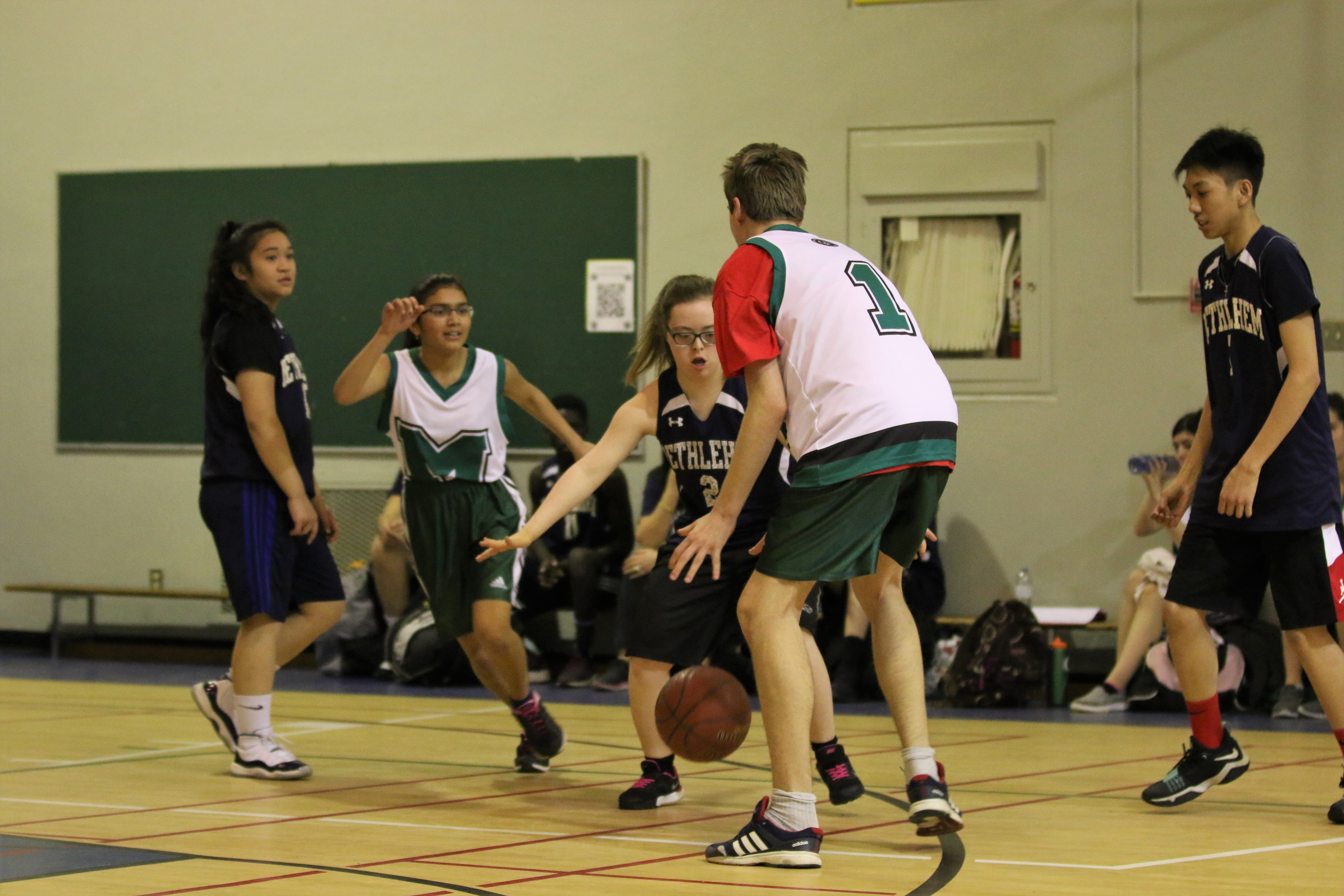 A action shot photo of a unified basketball game. Five athletes can be seen playing basketball on the court. One athlete is dribbling the ball while another is trying to defend him. A action shot photo of a unified basketball game. Five athletes can be seen playing basketball on the court. One athlete is dribbling the ball while another is trying to defend him.