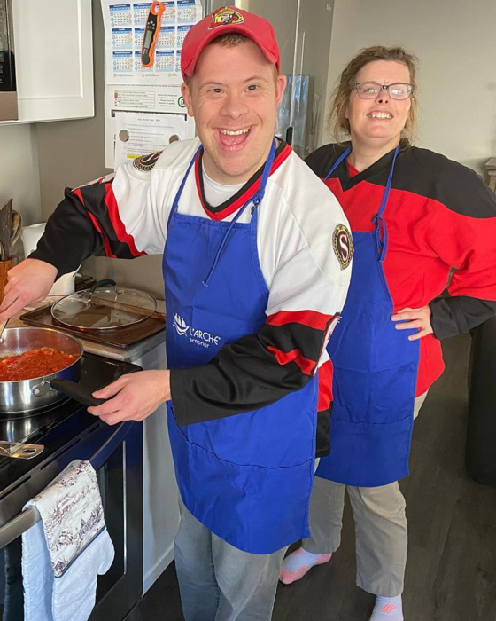 Une personne portant un chapeau rouge et un tablier bleu remue une casserole de sauce tomate tandis qu'une autre personne vêtue d'une chemise rouge et d'un tablier bleu observe.