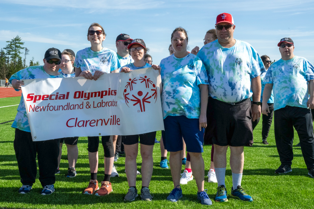 A group photo of the Clarenville Allstars Community Club. Everyone in the photo is wearing a blue tie-dye shirt. Three athletes are holding up a banner that says 