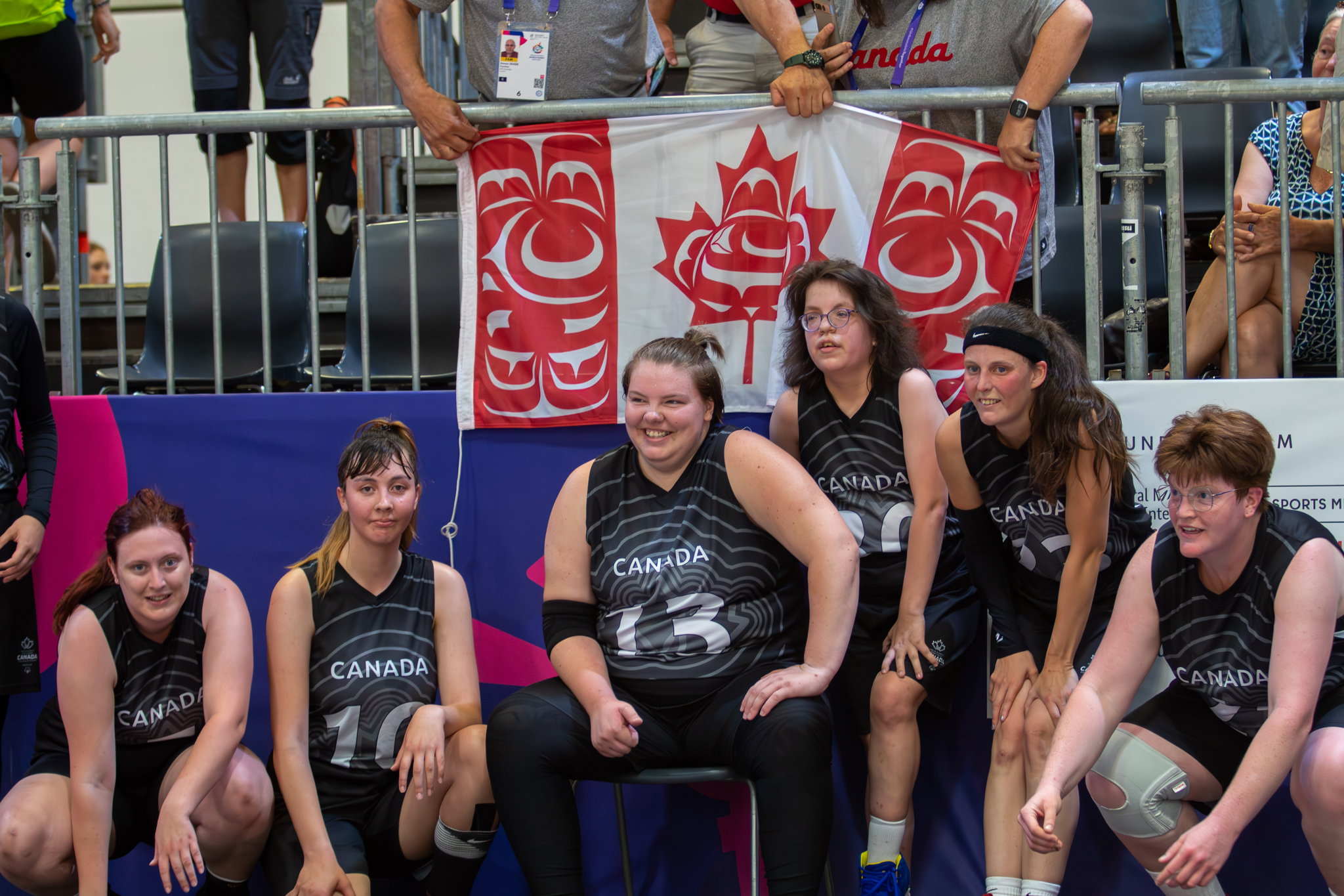 Six Special Olympics Saskatchewan athletes are sitting on the sidelines waiting for their turn to play. They are wearing black Canada jerseys with a different number on each of them. Supporters in the stands behind them are holding a Canadian flag.