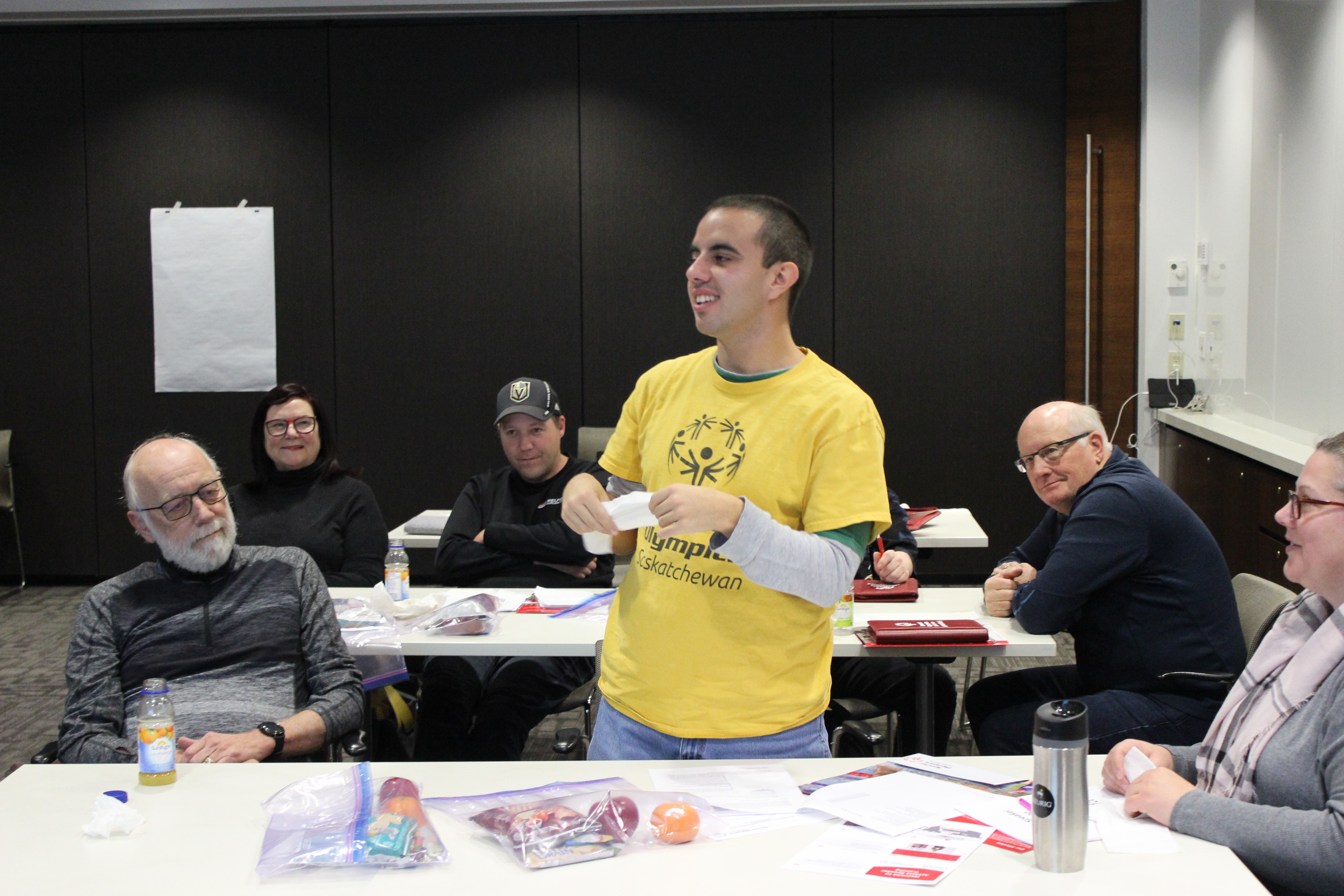 A conference room with three rectangular tables. People can be seen sitting at these three tables. One individual, in the middle of the image, in a yellow shirt, is standing up and presenting something to his peers. Everyone around him is listening intently. A conference room with three rectangular tables. People can be seen sitting at these three tables. One individual, in the middle of the image, in a yellow shirt, is standing up and presenting something to his peers. Everyone around him is listening intently.