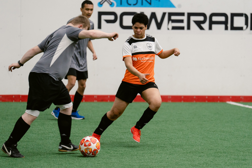 Three athletes playing soccer in an indoor soccer field. One athlete is wearing an orange jersey while the other two athletes are both in grey jerseys.