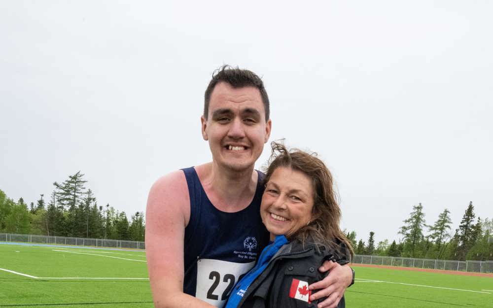 A picture of an athlete with their coach. The athlete has his arms around his coach like a hug. They are both standing and smiling for the camera.