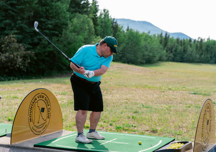 A golf player, wearing a bright blue golf shirt and black shorts with a green baseball cap, is taking his swing at the start of a golf game. Behind him you can see a forest with tall trees and mountains in the background.
