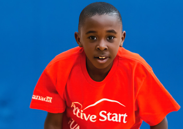 Young boy kneeling on a yellow bench. He is looking into the camera and wearing a red shirt that says ACTIVE START. 