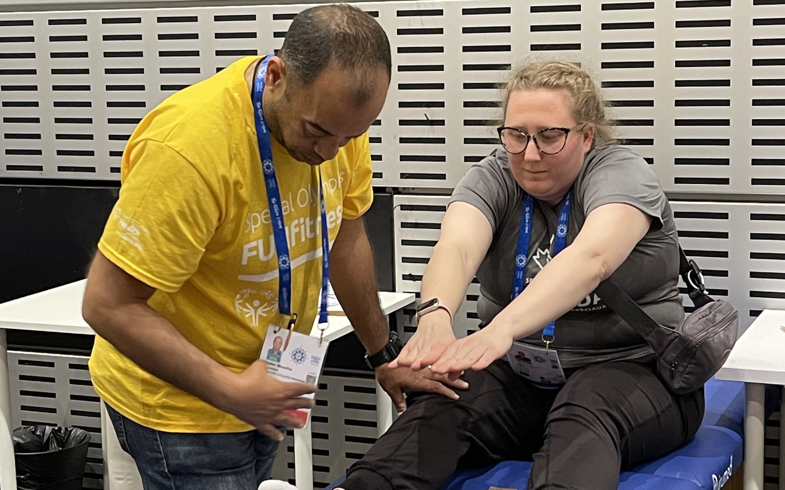 Young blond woman with glasses sitting on a blue medical bed. She is wearing dark pants and a grey shirt and stretching to reach her toes. A man in a yellow shirt is beside her bending down to check her flexibility. 