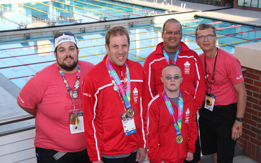 Bill Smith and teammates standing in-front of a swimming pool, showing off their medals. 