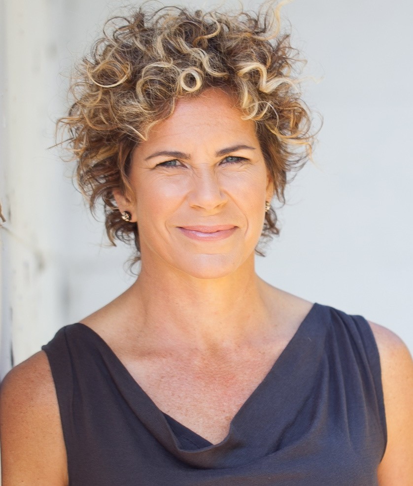 Headshot of a woman with curly blond short hair wearing a blue sleeveless blouse. Headshot of a woman with curly blond short hair wearing a blue sleeveless blouse.