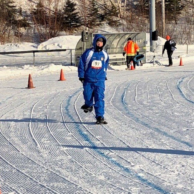 A photo of a snowshoeing athlete participating in his match. He is running around a curve that has been outlined with orange pylons. It is the winter and the athlete is wearing a blue snowsuit with the number "97" pinned to his coat.