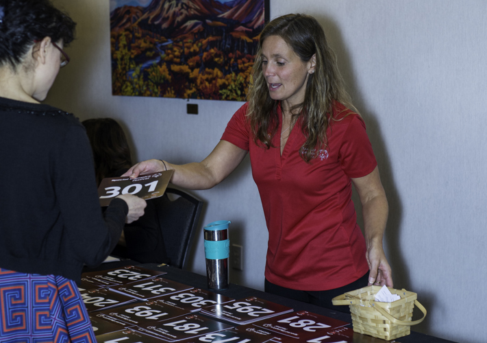 An event volunteer, in a red Special Olympics Yukon t-shirt, is giving out a number to an athlete. This interaction is occurring over a table lined up with more numbers.