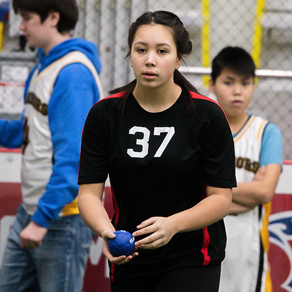 A close up image of a bocce player. She has her hair pulled back in a ponytail and is wearing a black jersey with the white number 37 on it. She is holding a blue bocce ball and is getting ready to throw it. She has a focused and serious look on her face.
