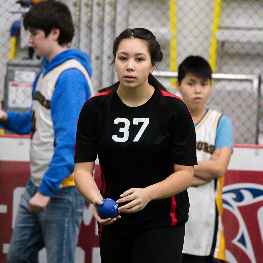 A close up image of a bocce player. She has her hair pulled back in a ponytail and is wearing a black jersey with the white number 37 on it. She is holding a blue bocce ball and is getting ready to throw it. She has a focused and serious look on her face.