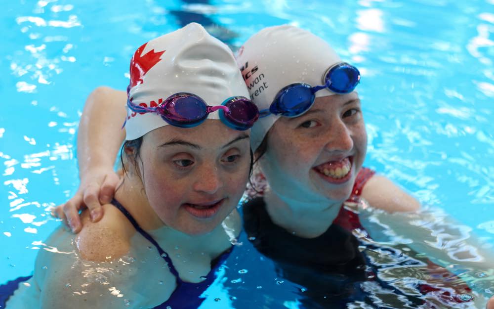 Two girls in a swimming pool. Both are wearing swim caps and have googles on their heads. One is smiling at the camera and the other is looking down at the pool.  