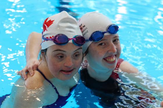 Two girls in a swimming pool. Both are wearing swim caps and have googles on their heads. One is smiling at the camera and the other is looking down at the pool.  