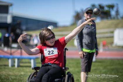 Person in a wheelchair with a red shirt and a number tag throwing a shot put Person in a wheelchair with a red shirt and a number tag throwing a shot put