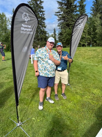 Hayden Metcalf and Hayden Metcalf’s father pose on a golf course beside Special Olympics British Columbia flags, with trees and sky in the background.