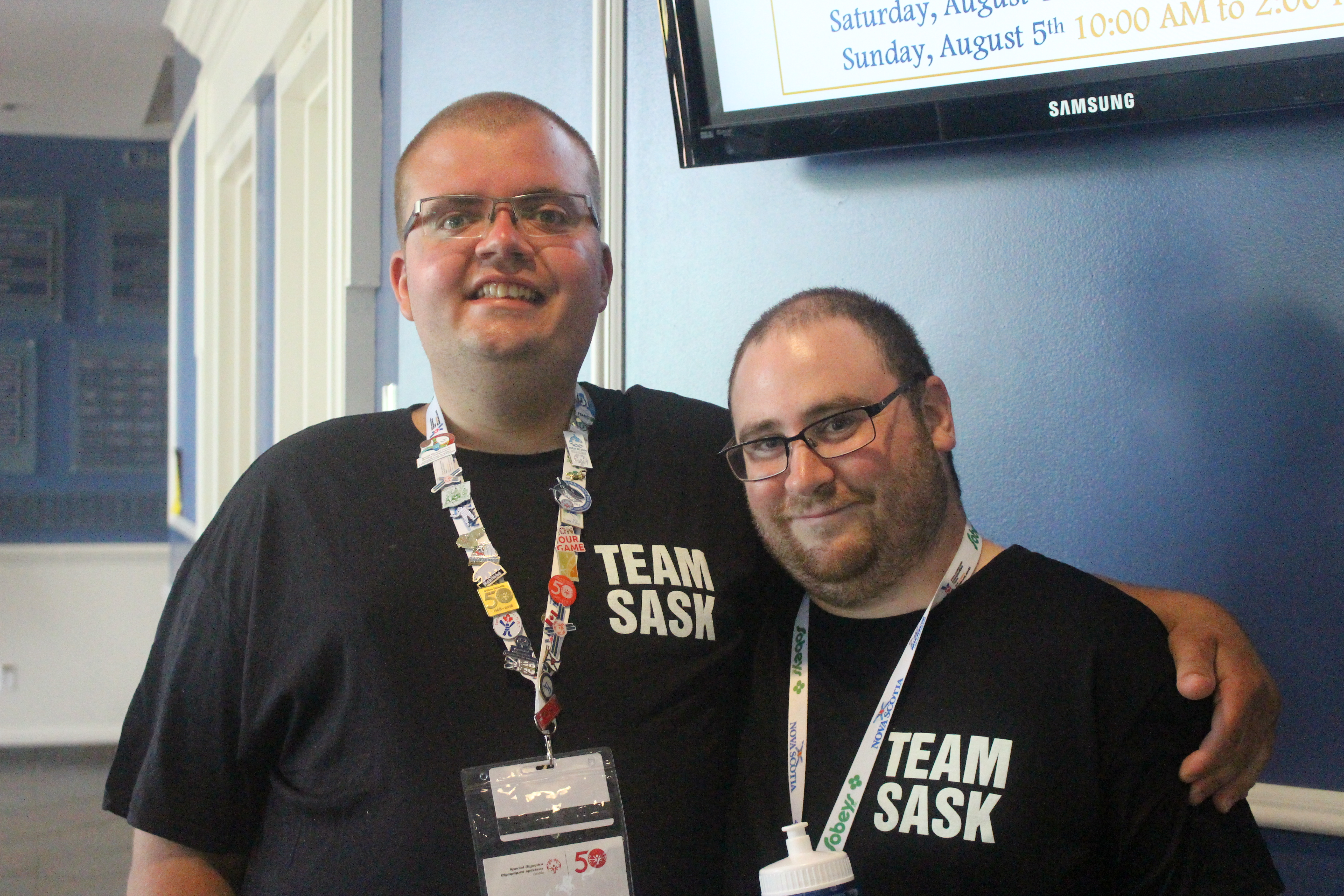 Two Special Olympics Saskatchewan athletes have their arms around and behind each other and are smiling for the camera. They are both wearing black 