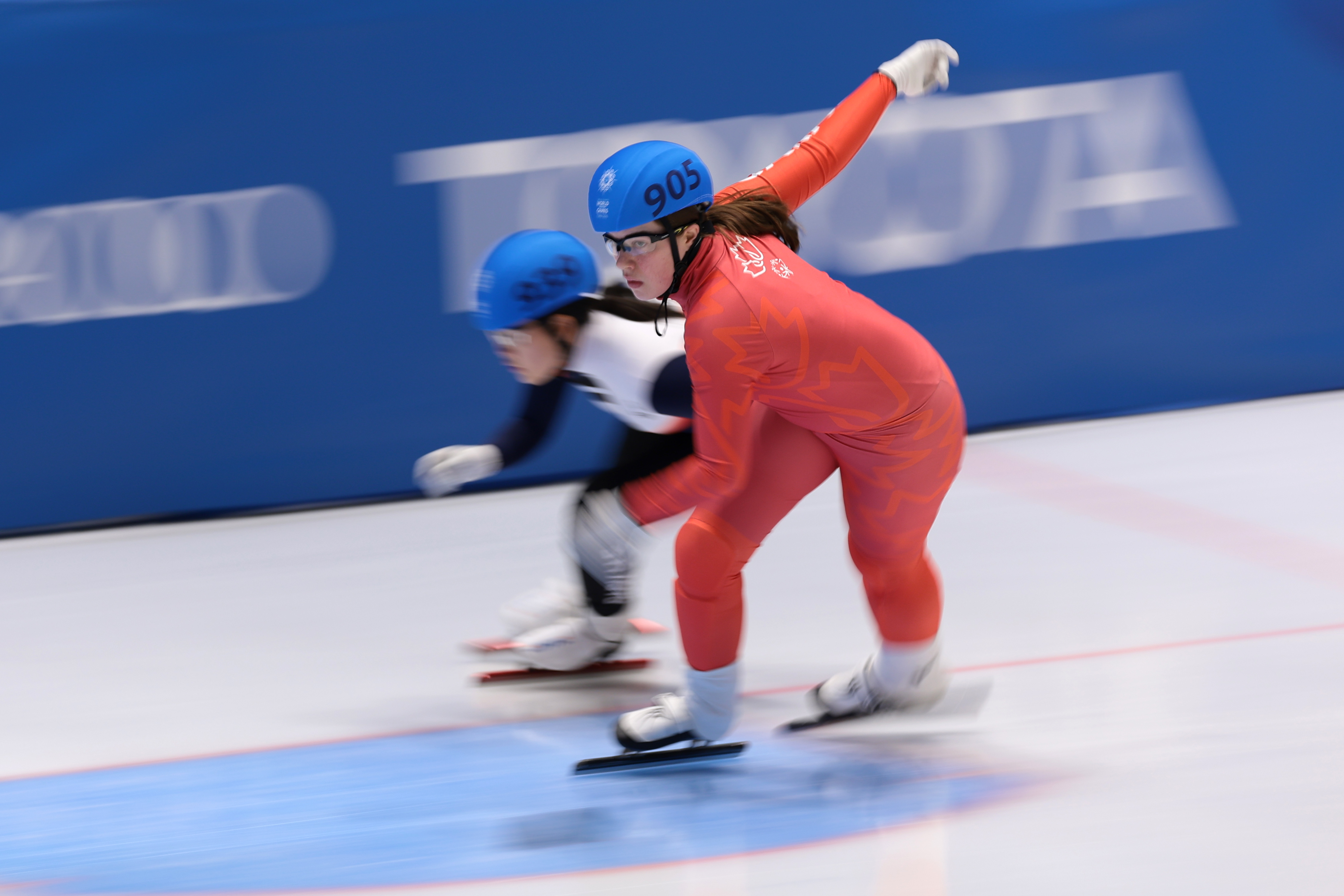 Canadian female speed skater in red skin suit. In the background is another speed skater in a black and white skin suit. Canadian female speed skater in red skin suit. In the background is another speed skater in a black and white skin suit.