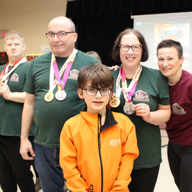 A group photo of multiple people. In the front is a young athlete wearing an orange Special Olympics Newfoundland & Labrador sweater. Behind him are three Special Olympics Newfoundland & Labrador athletes with medals hanging around their necks, as well as a volunteer.