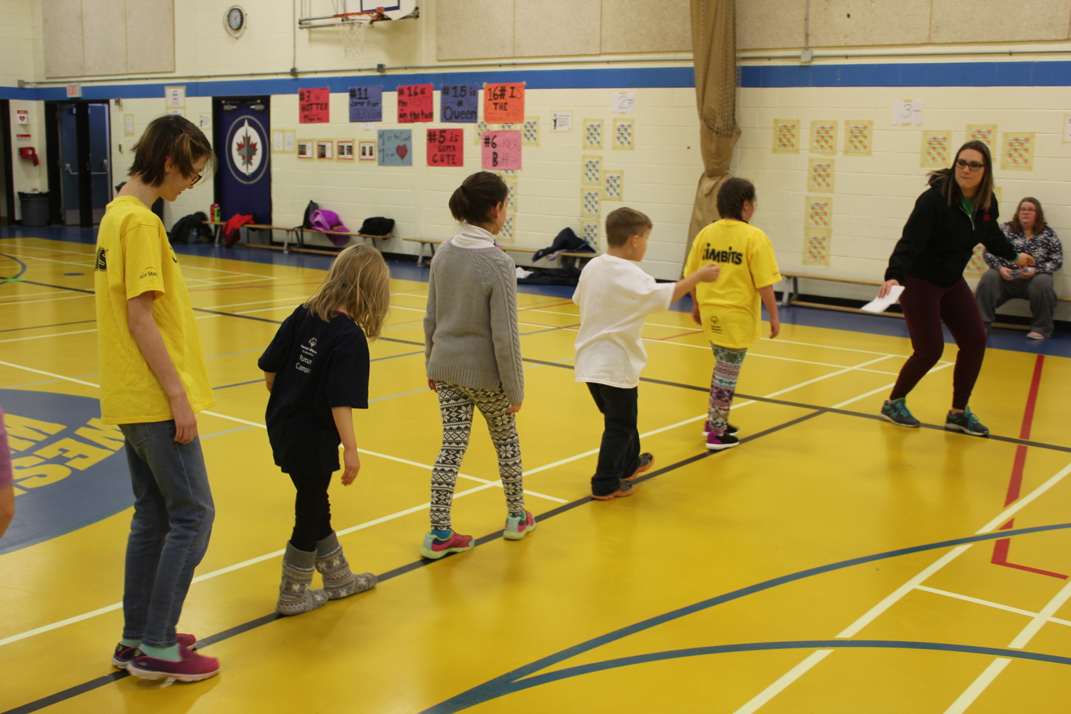 Five children in a line following their instructor. Everyone is in a gymnasium and the kids are following the lines on the floor. We can only see the children's backs as they are facing away from the camera.