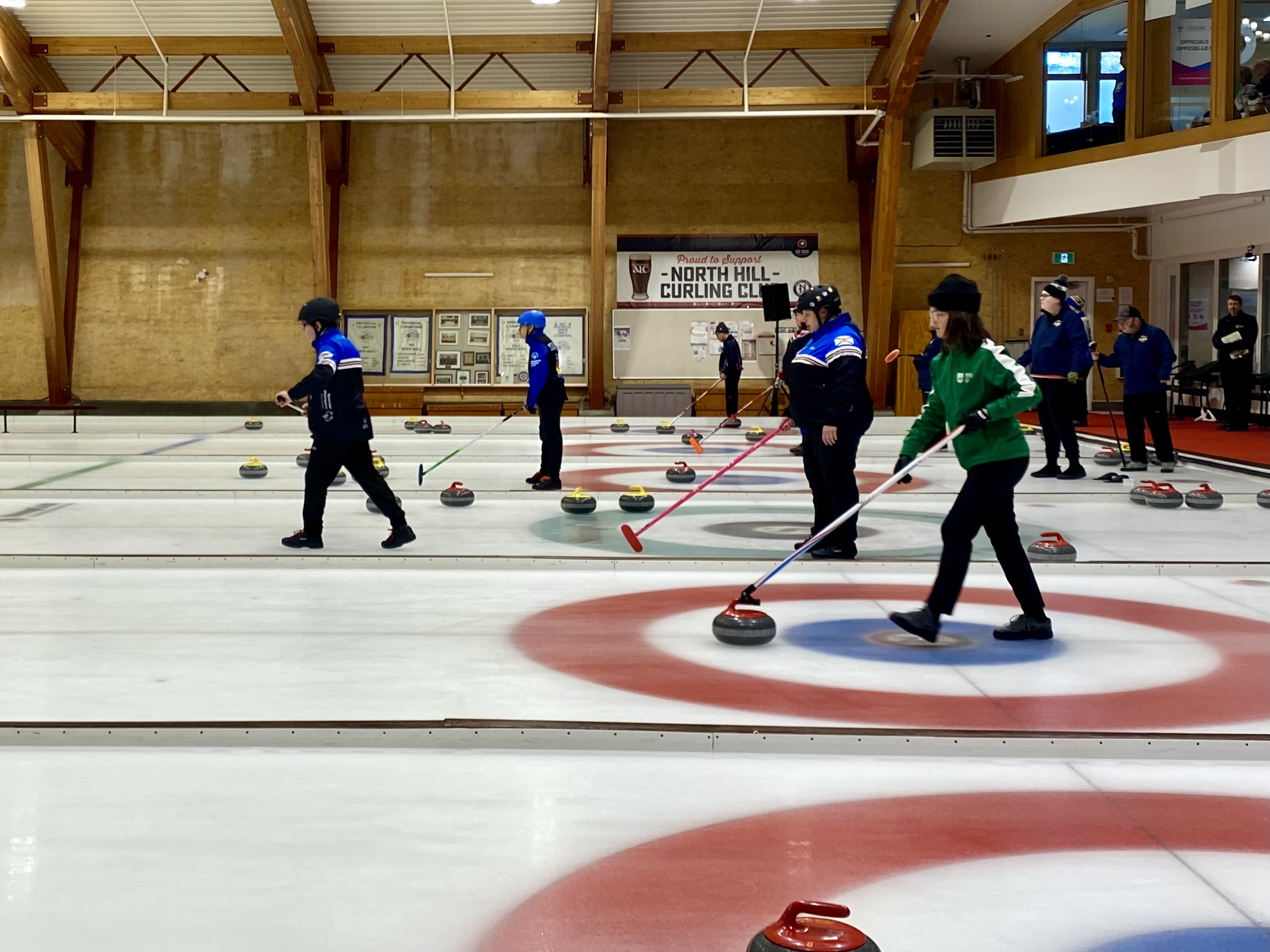 A wide shot action photo of curling games in play. Multiple curling athletes can be seen with their curling sticks, getting ready to move the curling stone across the ice. A wide shot action photo of curling games in play. Multiple curling athletes can be seen with their curling sticks, getting ready to move the curling stone across the ice.