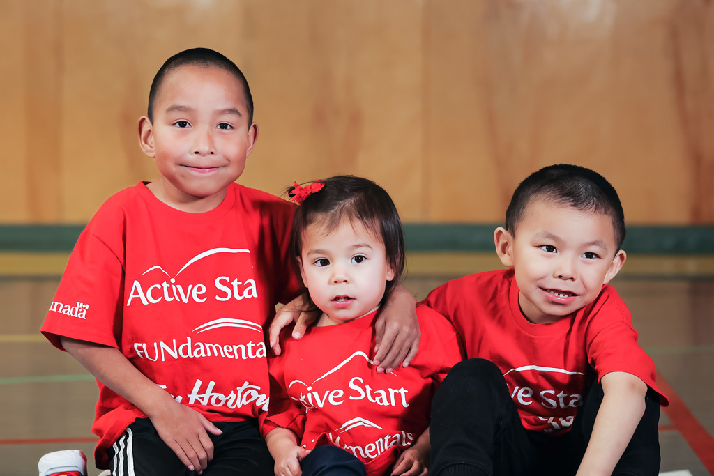 Trois jeunes - deux garçons et une fille - assis par terre. Ils semblent avoir entre 5 et 7 ans. Tous portent des t-shirts rouges sur lesquels est écrit ACTIVE START. Ils sourient tous.