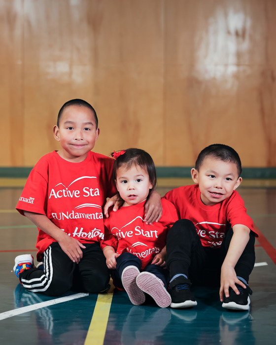 Three young people - two boys and a girl - sitting on the floor. They appear to be about 5-7 years old. All are wearing red shirts that say ACTIVE START. All of them are smiling. 