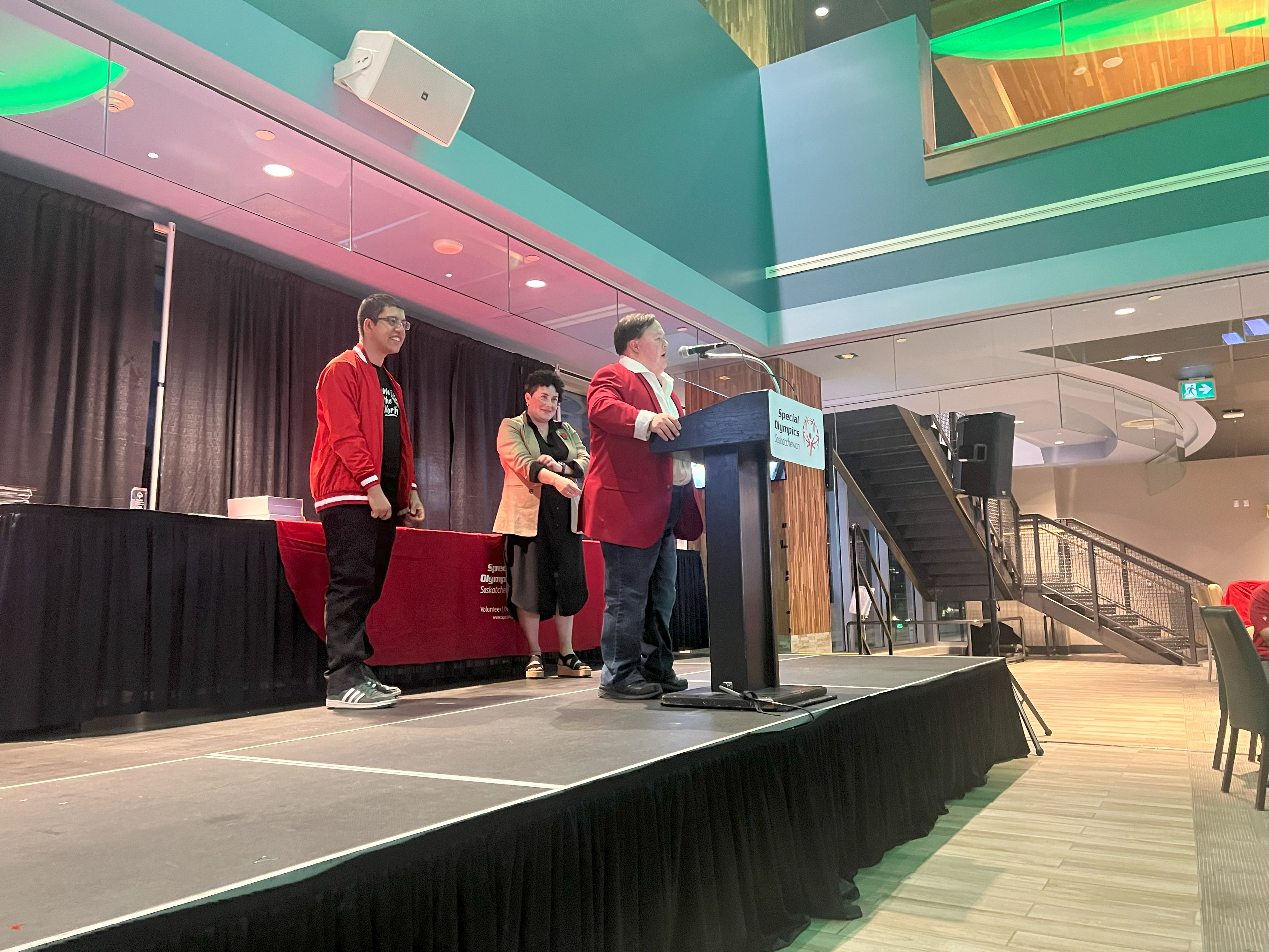 Three individuals are standing on a stage. The angle is taken from off stage looking up at them diagonally. Two individuals are standing back while one is at the podium giving a speech. Three individuals are standing on a stage. The angle is taken from off stage looking up at them diagonally. Two individuals are standing back while one is at the podium giving a speech.