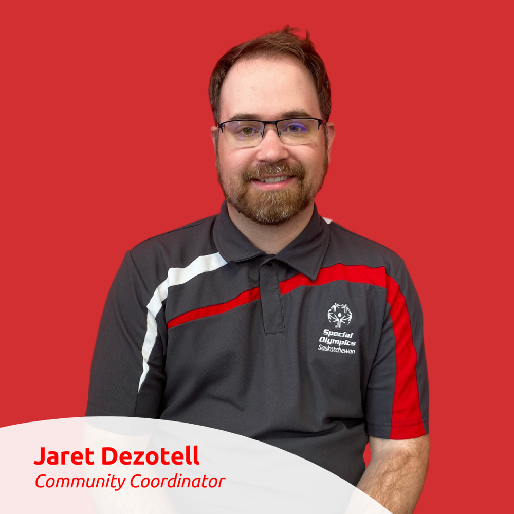 Portrait Photo of Jaret Dezotell. He is standing against a red wall and smiling for the camera.