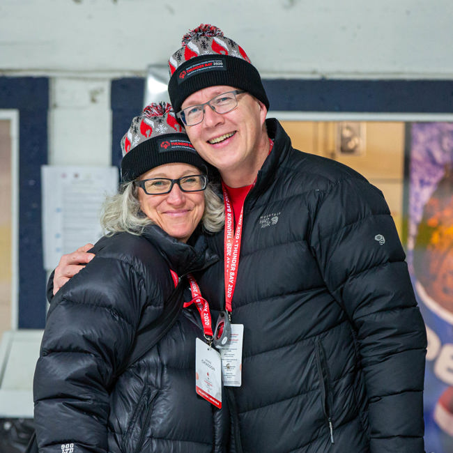 Two people in black winter jackets and Thunder Bay 2020 toques smile at the camera 