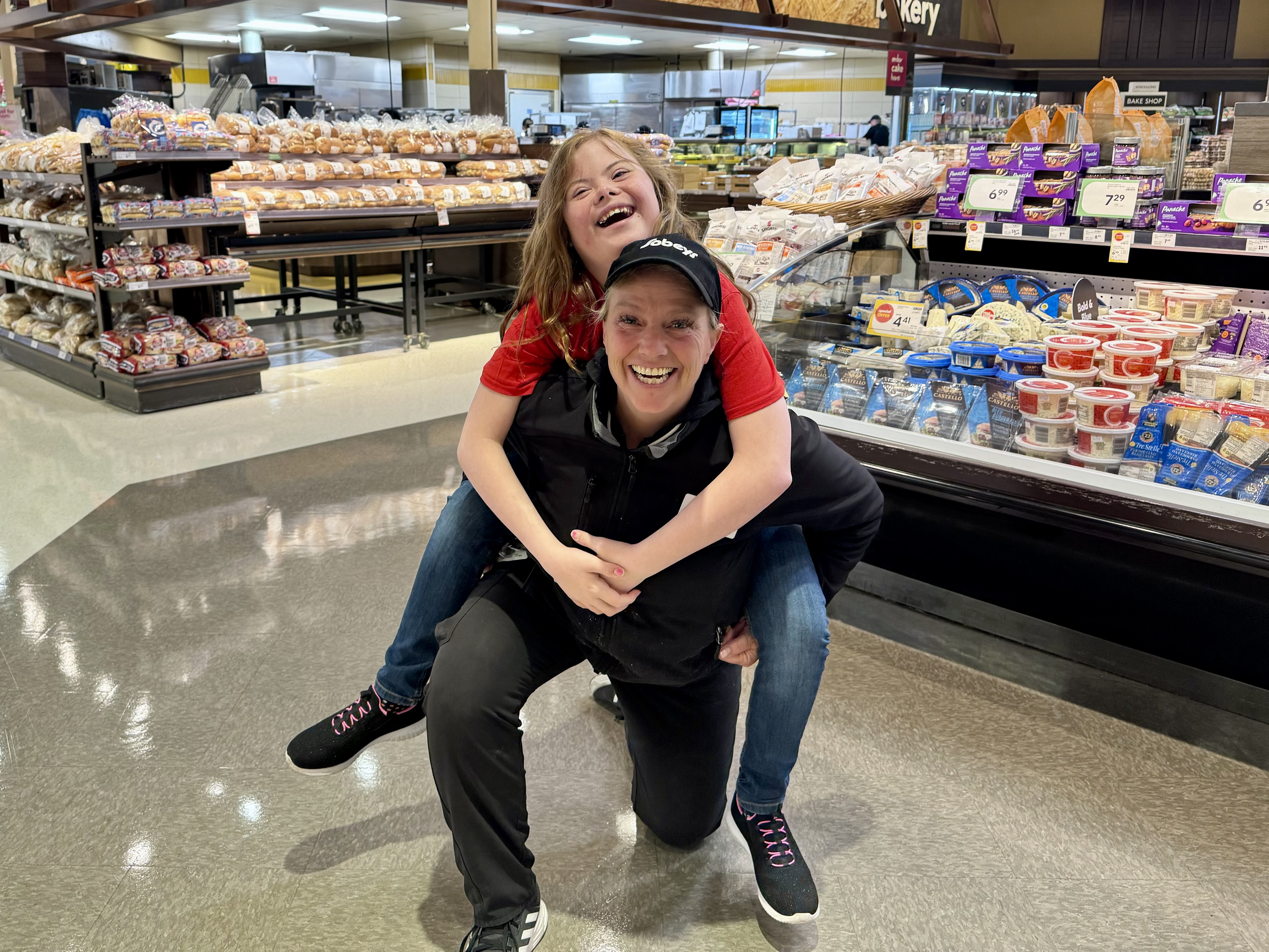 A young athlete is smiling and piggybacking a Sobeys employee. The Sobeys employee is also smiling for the camera. Behind them are grocery aisles stocked with different food assortments.