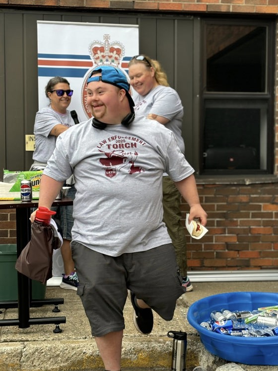 An athlete is walking towards the camera, but looking to the left. He is smiling happily with a hot dog in one hand and his water bottle in another. He is wearing a grey Law Enforcement Torch Run t-shirt and a blue baseball cap, facing backwards.