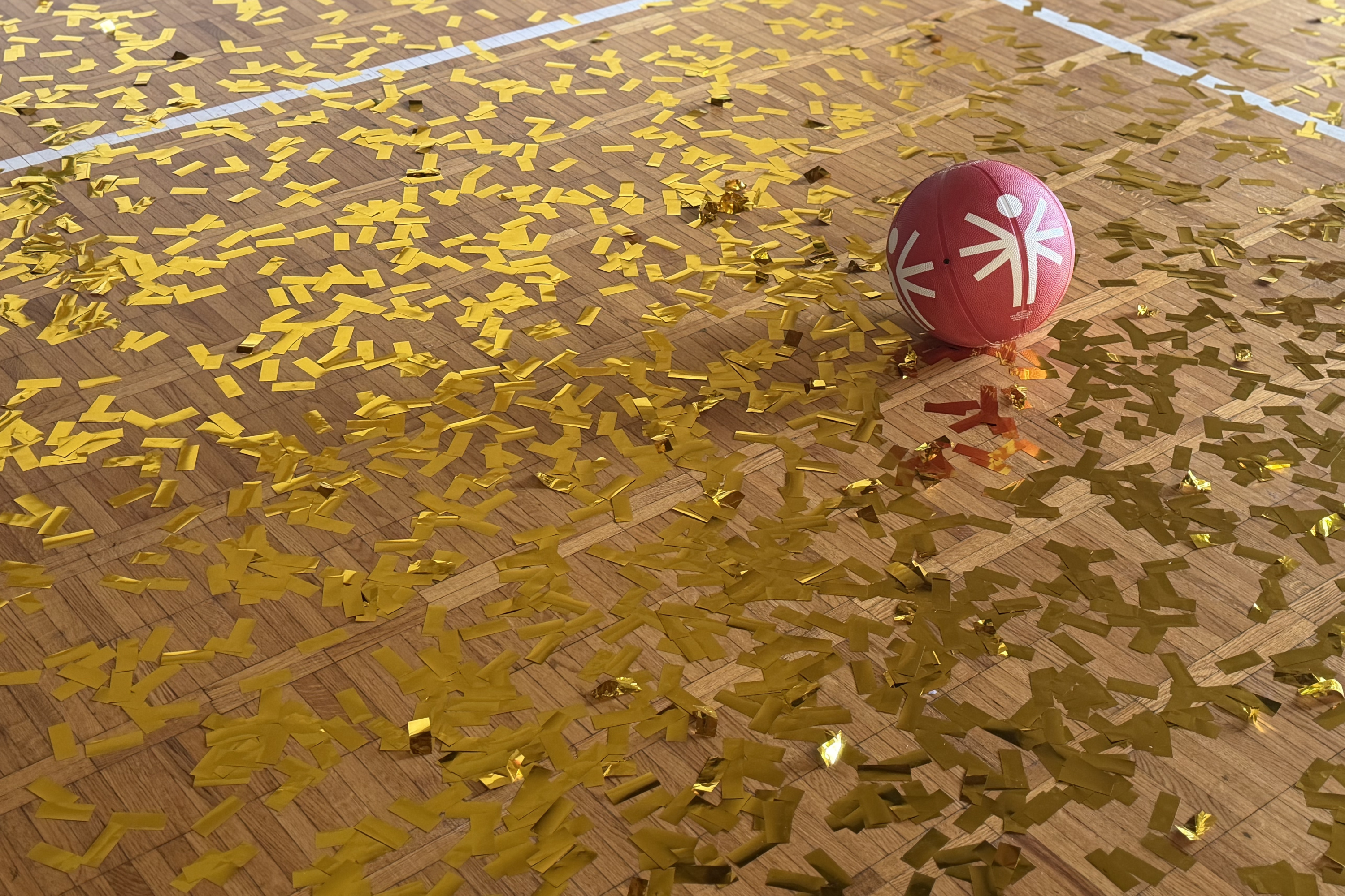 Gold confetti spread across a gym floor, a red basketball sits on top. Gold confetti spread across a gym floor, a red basketball sits on top.