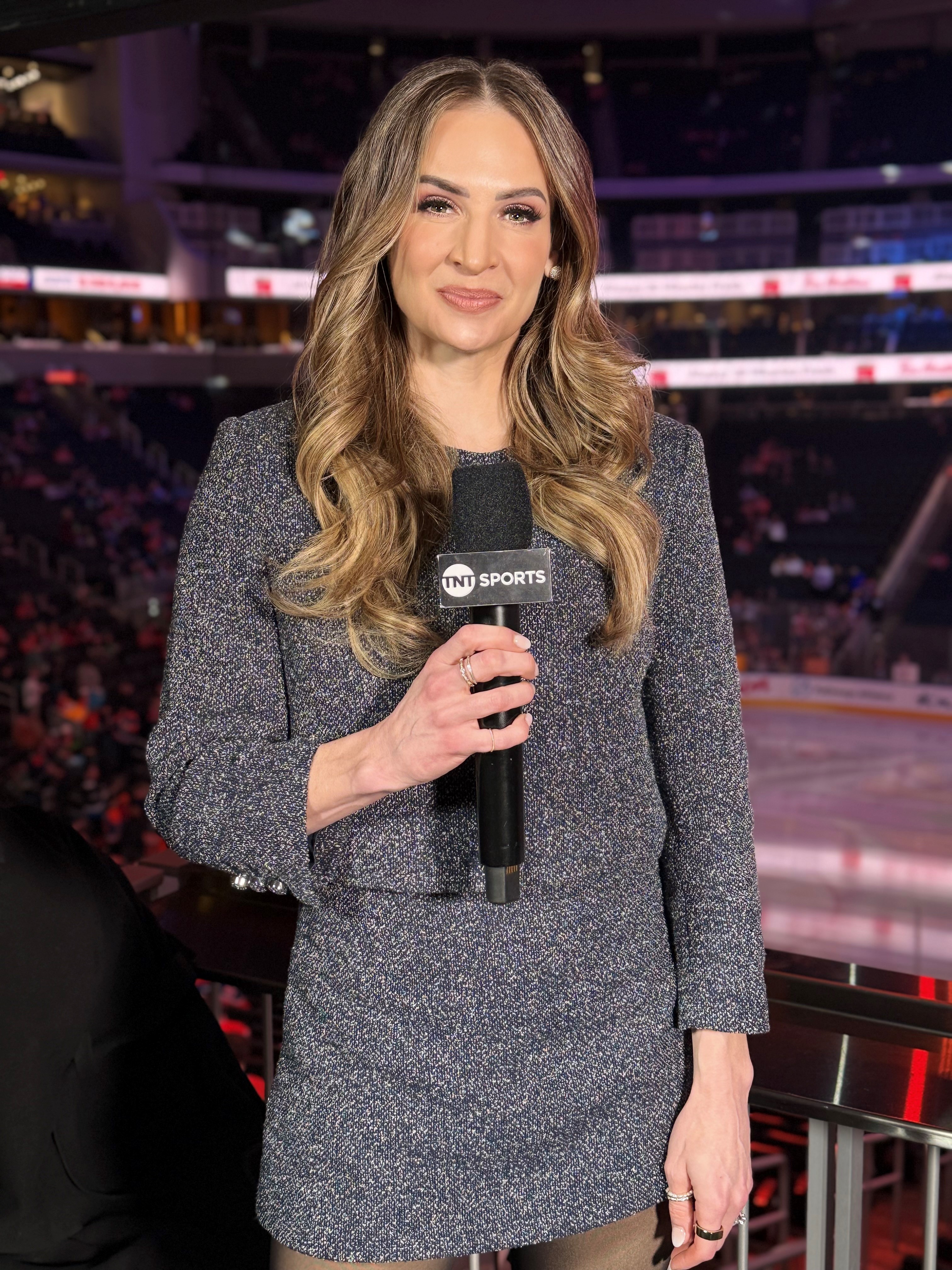 Headshot of a woman with curly long hair wearing a grey dress, holding a microphoe and standing in a hockey arena. Headshot of a woman with curly long hair wearing a grey dress, holding a microphoe and standing in a hockey arena.