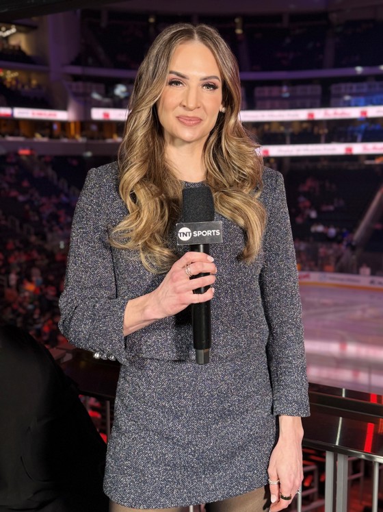 Gros plan sur une femme aux longs cheveux bouclés, vêtue d'une robe grise, tenant un micro et debout dans une patinoire de hockey.