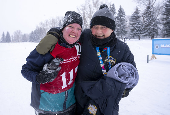 Two people with their arms around each other, smiling into the camera. One is a woman in a red pinny and one is a man with a black jacket. The background is covered in snow. 