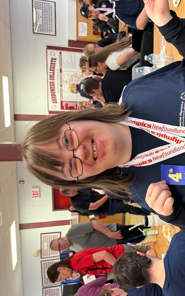 A Special Olympics Newfoundland & Labrador athlete is smiling for the camera and posing with her awards. She has two bronze medals hanging around her neck and she's holding a blue 4th place ribbon.