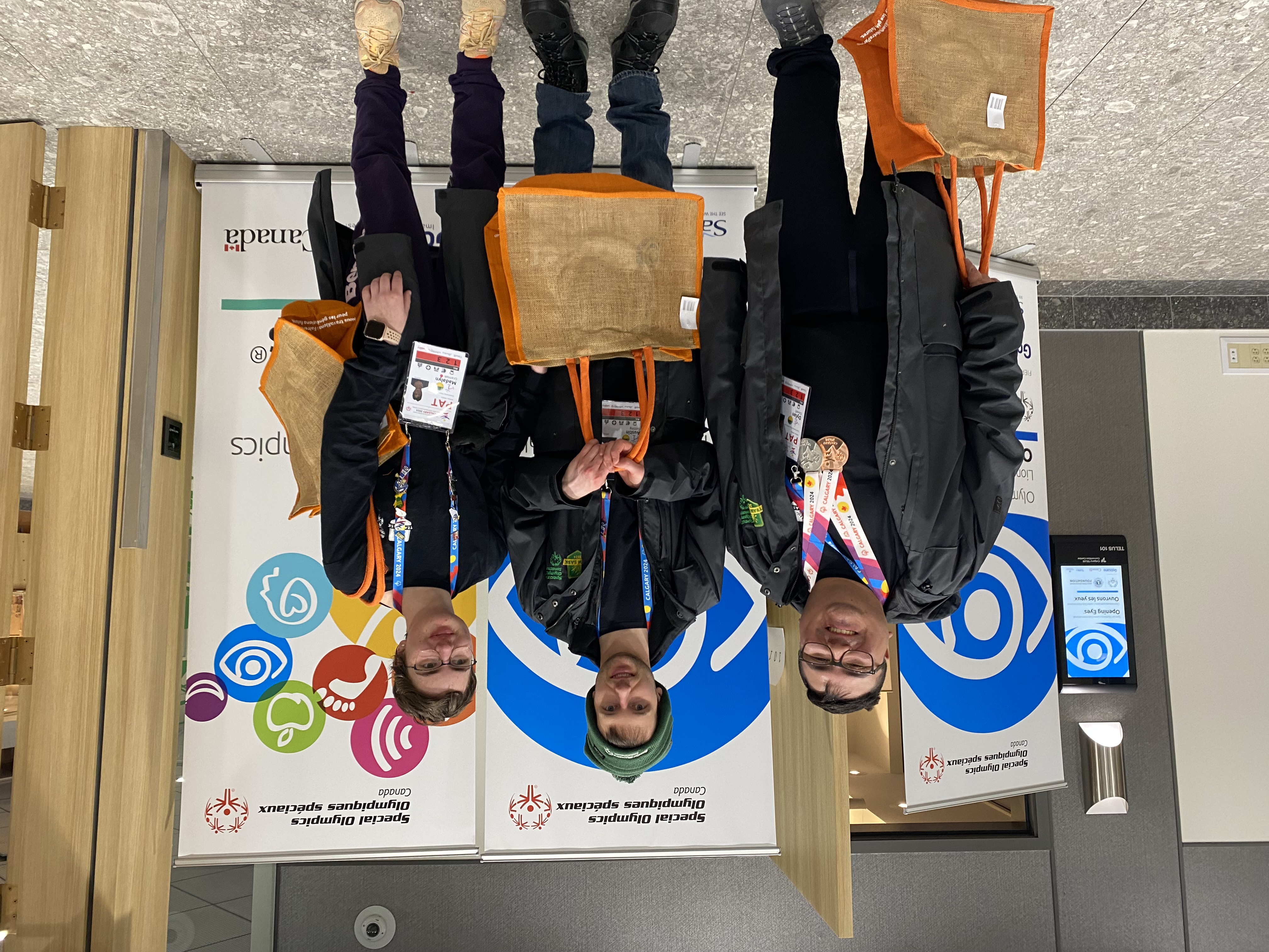 Three athletes are standing in front of Healthy Athletes banners and smiling for the camera. They are holding orange tote bags, and wearing all black with colourful lanyards around their necks.